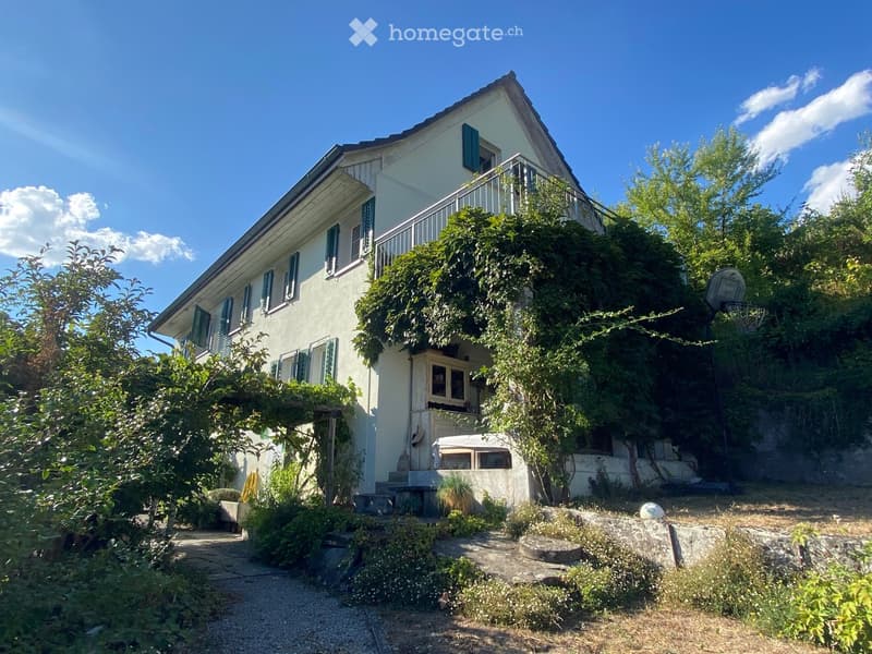 Two-story house with light-colored exterior, green shutters, and a balcony on the upper level. The house is surrounded by lush greenery and a stone pathway leads to the entrance.