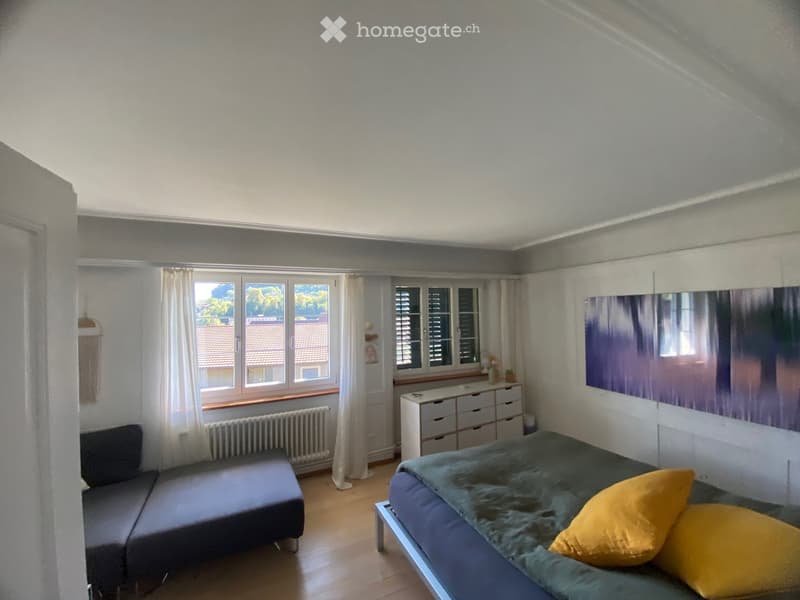 Bright bedroom with a double bed, sofa, dresser, large mirror, and large windows with curtains.