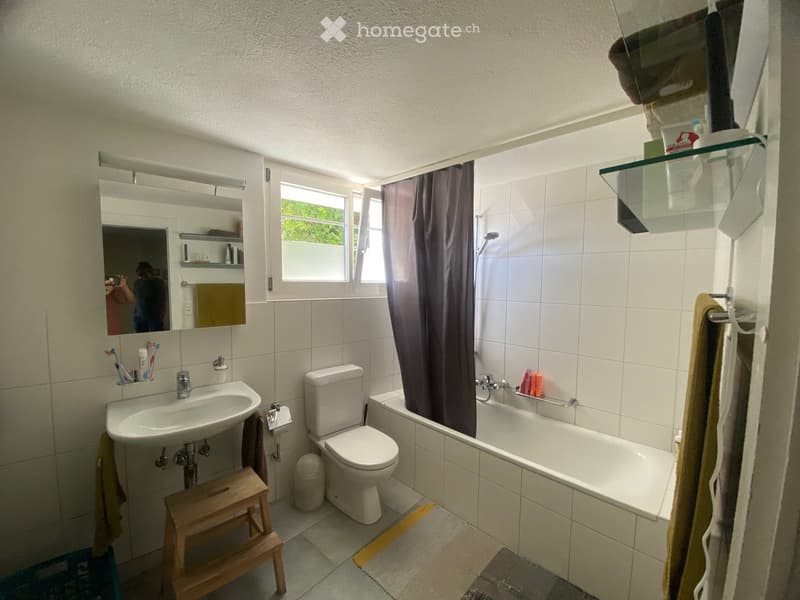 Bathroom with a sink, toilet, bathtub, mirror, and a small wooden stool. The room has white tiled walls and a window with a dark curtain.