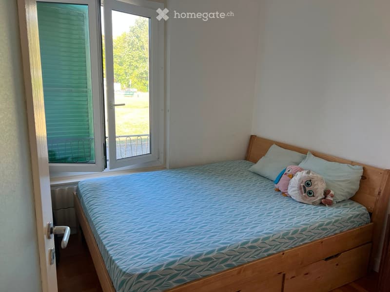 A small bedroom with a single bed, light blue bedding, and a stuffed animal on the bed. The room has a window with blinds and a radiator below it.