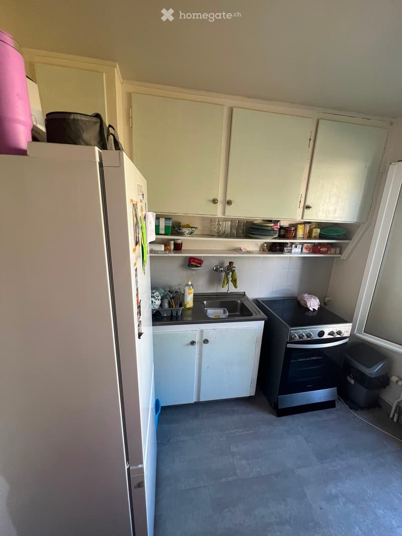 A compact kitchen with white cabinets, a stainless steel sink, a black oven, and a refrigerator. The countertops hold various kitchen items, and the floor is tiled.