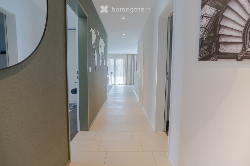 Modern hallway with light-colored tiled flooring, white walls, and recessed ceiling lighting. Features a decorative world map mural on one wall and a framed staircase photograph on another.