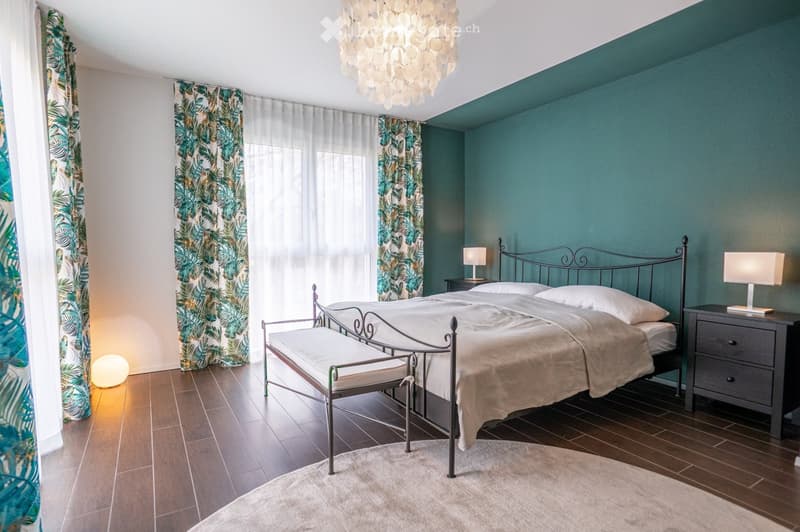 Cozy bedroom with teal accent wall, wooden floor, large bed with metal frame, nightstands with lamps, bench at foot of bed, and decorative rug.