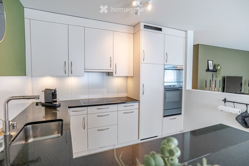 Modern kitchen with white cabinets, stainless steel sink, coffee maker, and built-in oven. Features a sleek black countertop and a small decorative display with candles and a mirror.