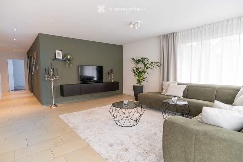Modern living room with a green accent wall, a dark wooden TV stand, a grey sofa with white cushions, a geometric glass coffee table, and a large plant in a pot.