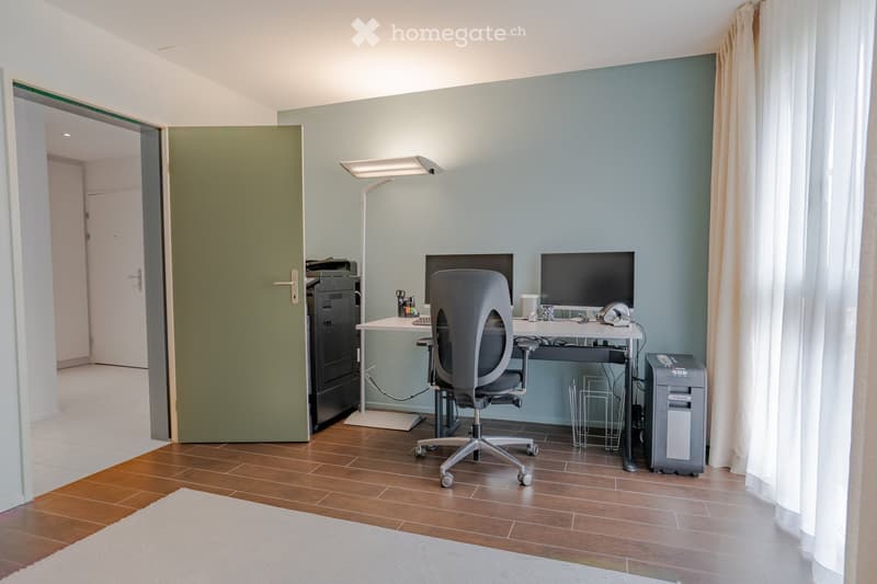 Modern office space with wooden flooring, a desk with a computer, a chair, a printer, and a lamp. A large window with curtains is on the right side, and a green door is visible on the left.