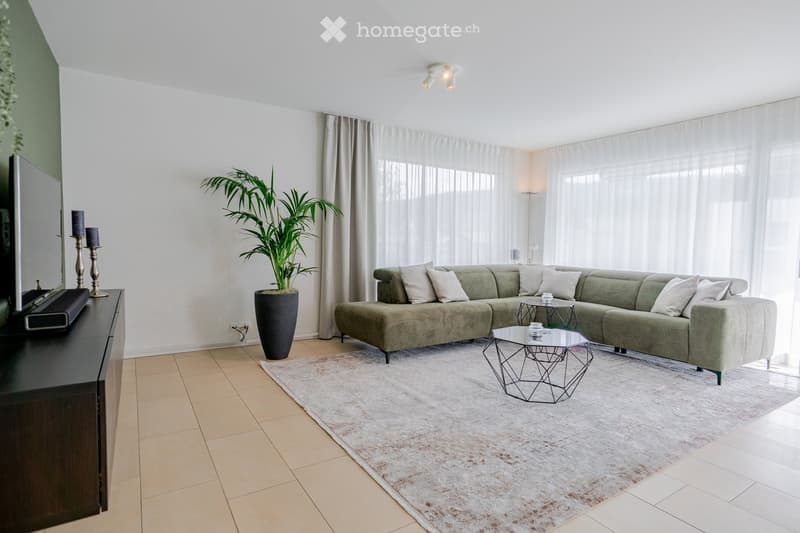 Modern living room with a green sectional sofa, coffee table, side table, television, candle holders, and a large plant in a pot. The room features large windows with sheer curtains and a light-colored rug on the floor.