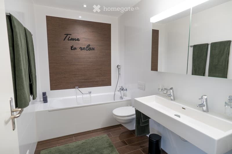 Modern bathroom with a bathtub, toilet, sink, large mirror, and decorative towel. Features a wooden accent wall with the phrase "Time to relax" and a green bath mat on the floor.
