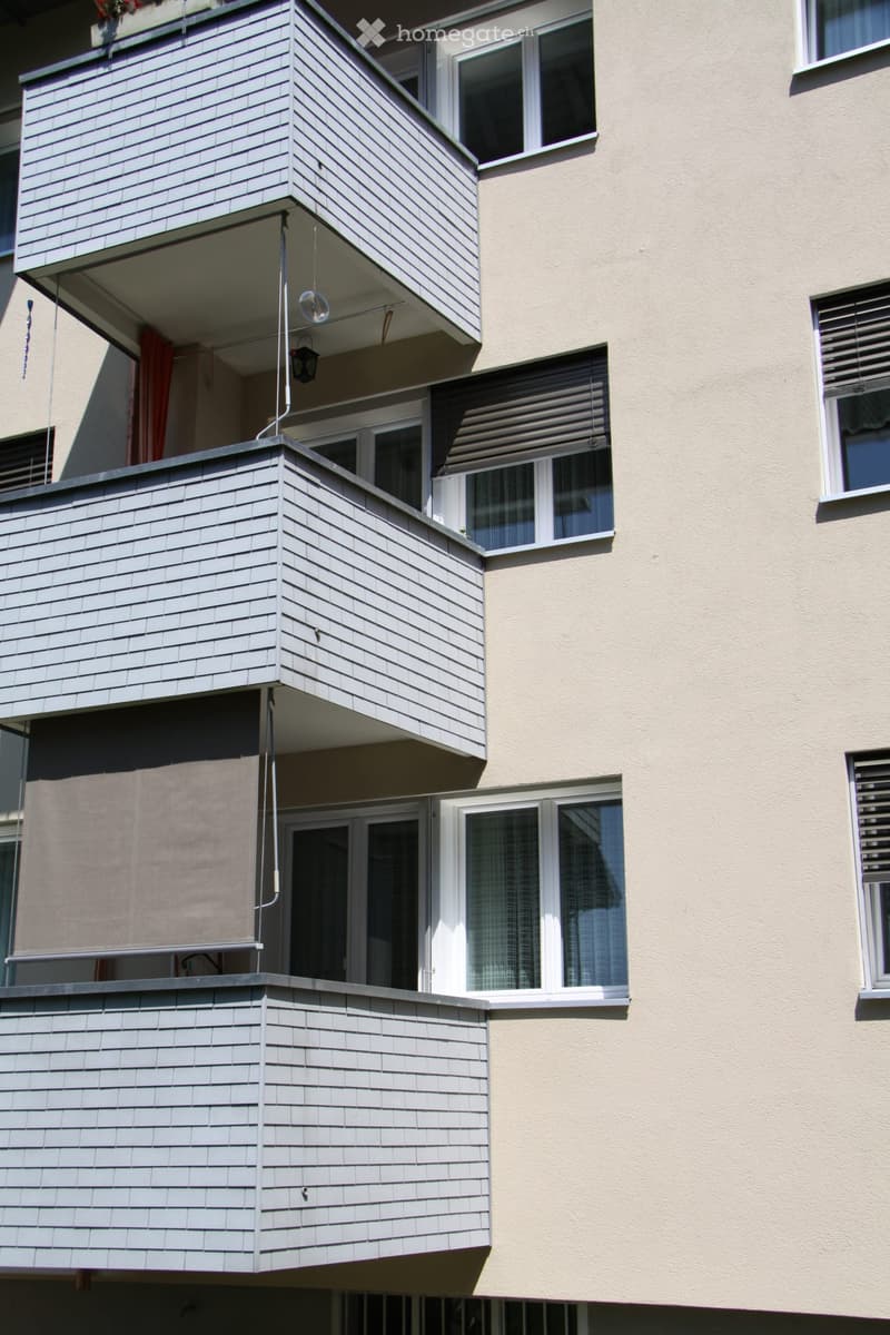 Balconies with grey wooden paneling and white railings, attached to a beige building with multiple windows and shutters.