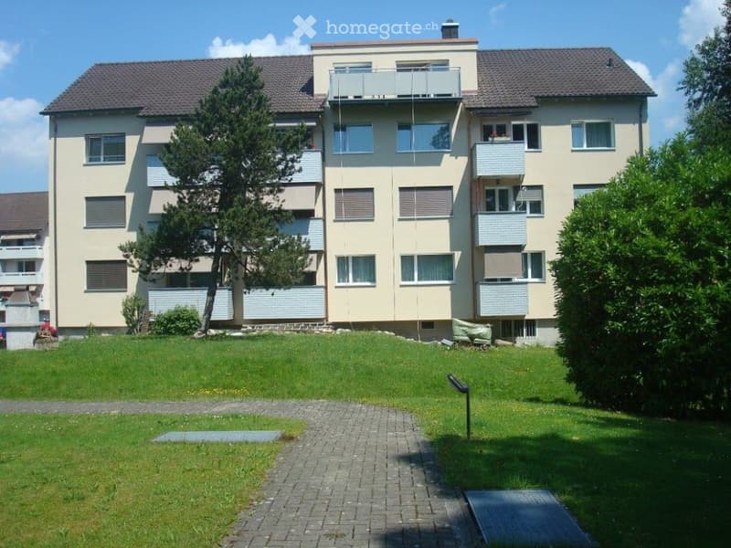 Multi-story residential building with balconies, surrounded by a well-maintained lawn and trees.