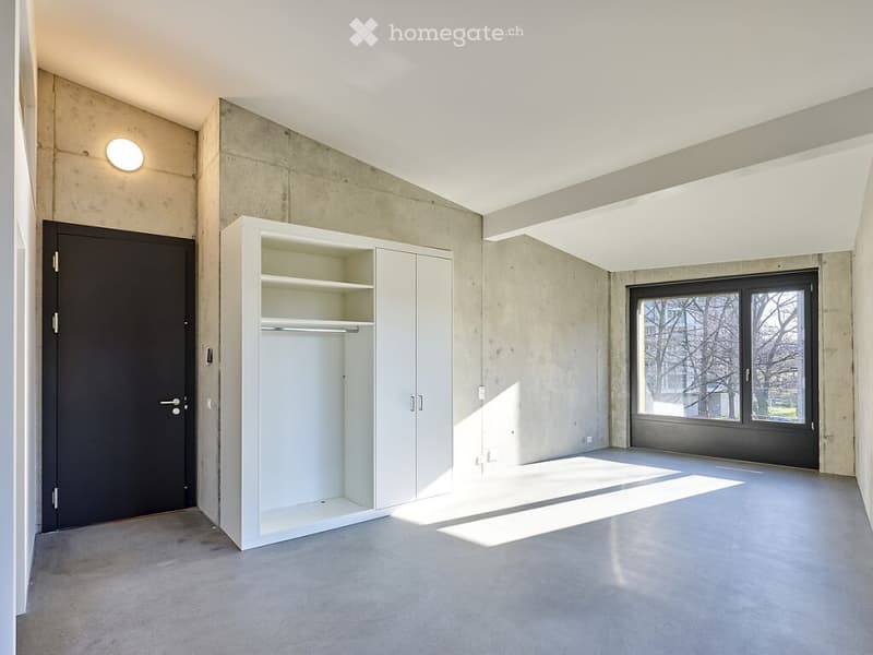 Empty room with concrete walls and floor, white built-in closet, large window, and a single light fixture on the wall.
