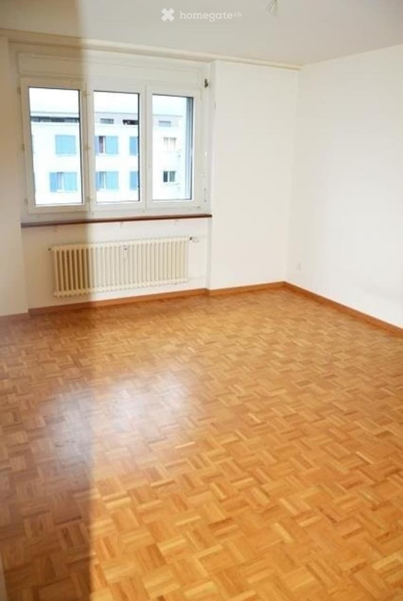 Empty room with wooden flooring, white walls, large windows, and a radiator below the windows.