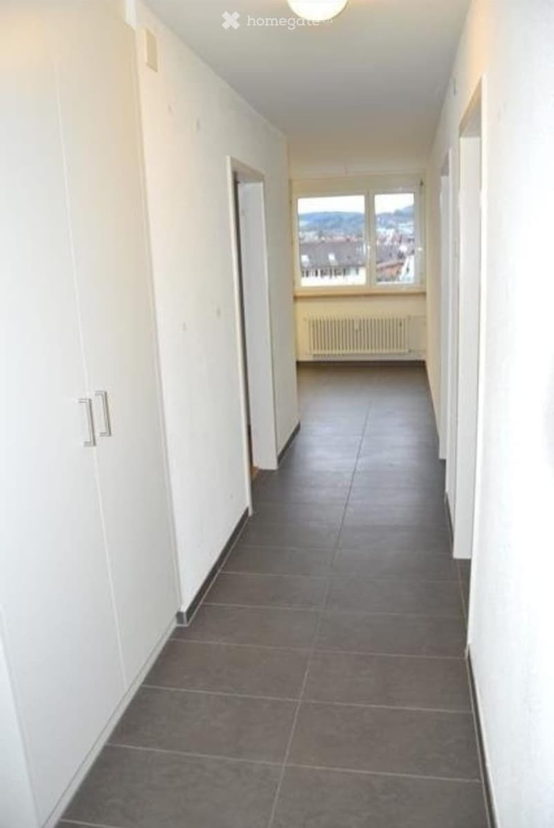 Empty hallway with tiled floor, white walls, and a view of a room with a radiator and large window at the end.