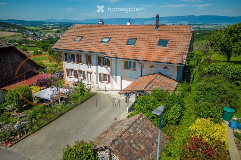 A two-story house with a red-tiled roof and white walls, surrounded by a well-maintained garden and outdoor seating area. The house features multiple windows, a front entrance, and is situated in a scenic countryside setting with distant hills.