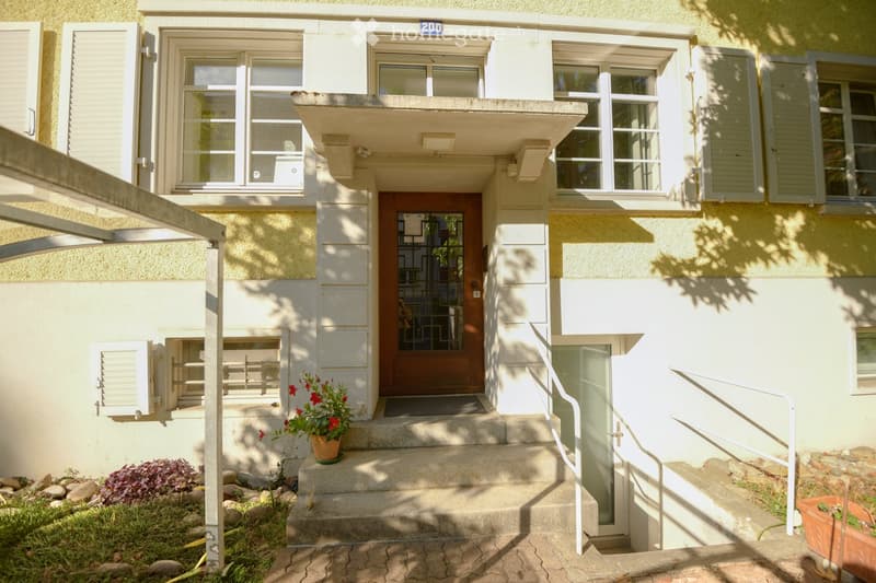 Two-story residential building with a balcony, entrance steps, and potted plants near the entrance.