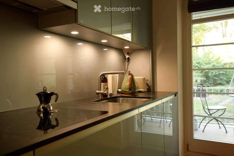 Modern kitchen with sleek, light-colored cabinets, a stainless steel sink, and a countertop with a coffee maker and toaster. A large window offers a view of the outdoor area.