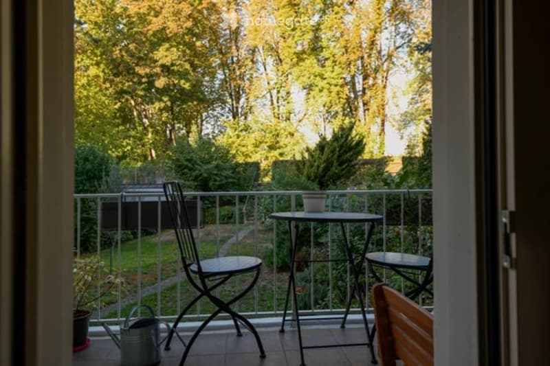 Balcony with outdoor furniture including a small table, chairs, and potted plants, overlooking a green garden area.