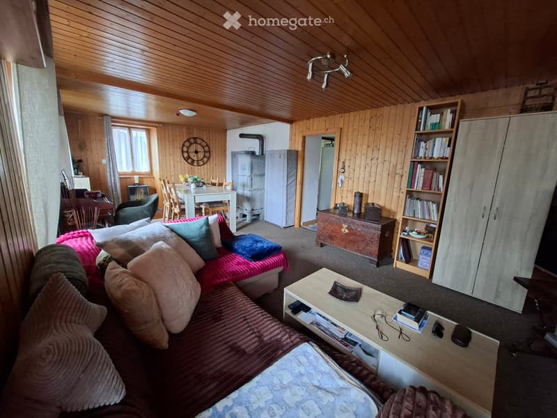 Cozy living room with wooden walls and ceiling, featuring a red sofa, coffee table, bookshelf, dining area with table and chairs, and a refrigerator in the background.