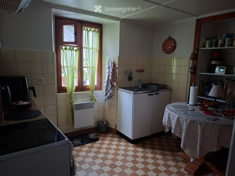 The kitchen features a checkered tile floor, white cabinets, a sink, a stove, and a small table with a tablecloth. There are various kitchen items on shelves and a window with green curtains.