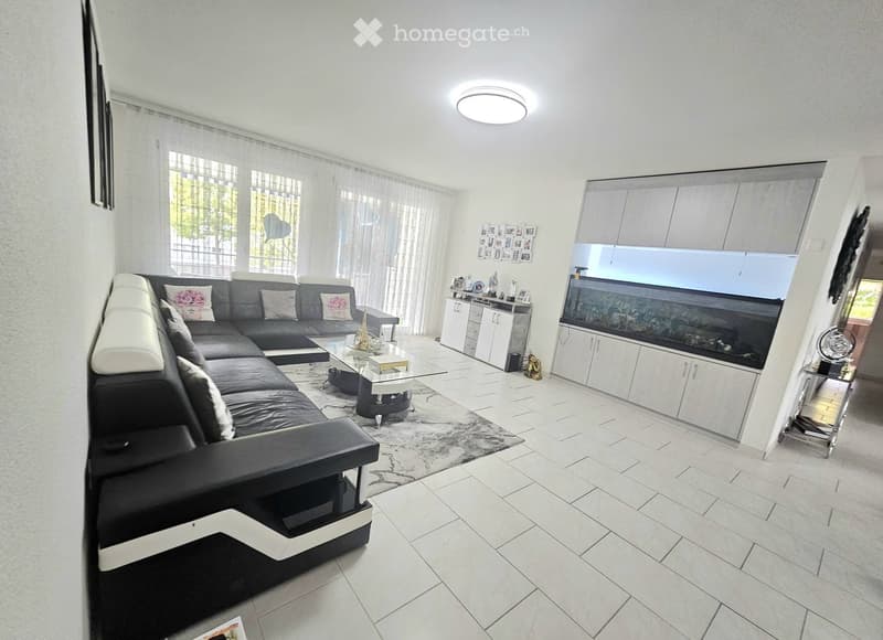 Modern living room with a black sectional sofa, glass coffee table, fireplace, and white cabinets. The room features large windows with sheer curtains and a tiled floor.