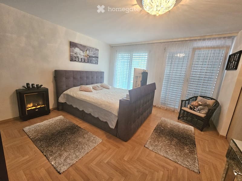 Cozy bedroom with a large upholstered bed, fireplace, wicker chair with stuffed animals, and large windows with curtains.