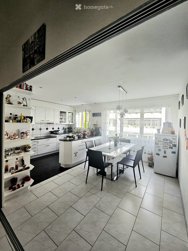 Modern kitchen with white cabinets, black countertops, and a central island. Features a dining table with chairs, a refrigerator, and various kitchen appliances and decor items.