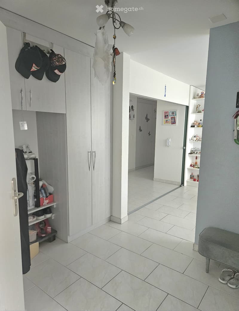 Bright hallway with tiled flooring, white walls, built-in storage units, and a small seating area with a grey ottoman and shoes placed nearby.