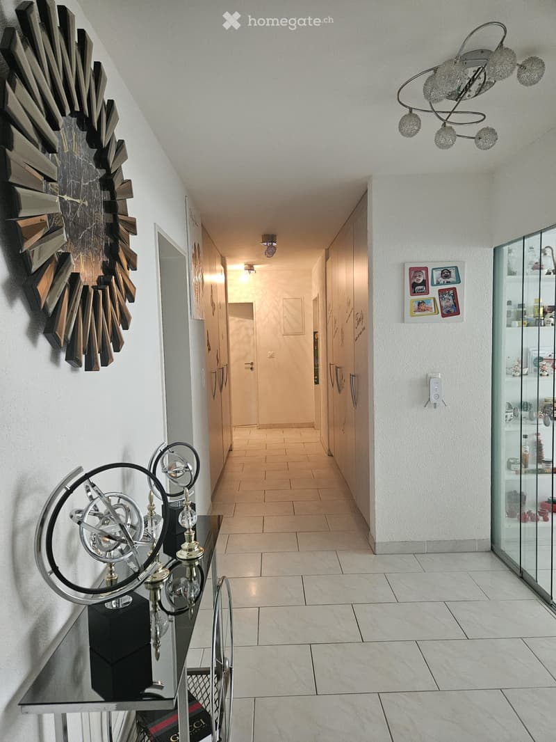 Modern hallway with light-colored tiles, glass display cabinet, decorative wall clock, and a small table with a chandelier-like sculpture on top.