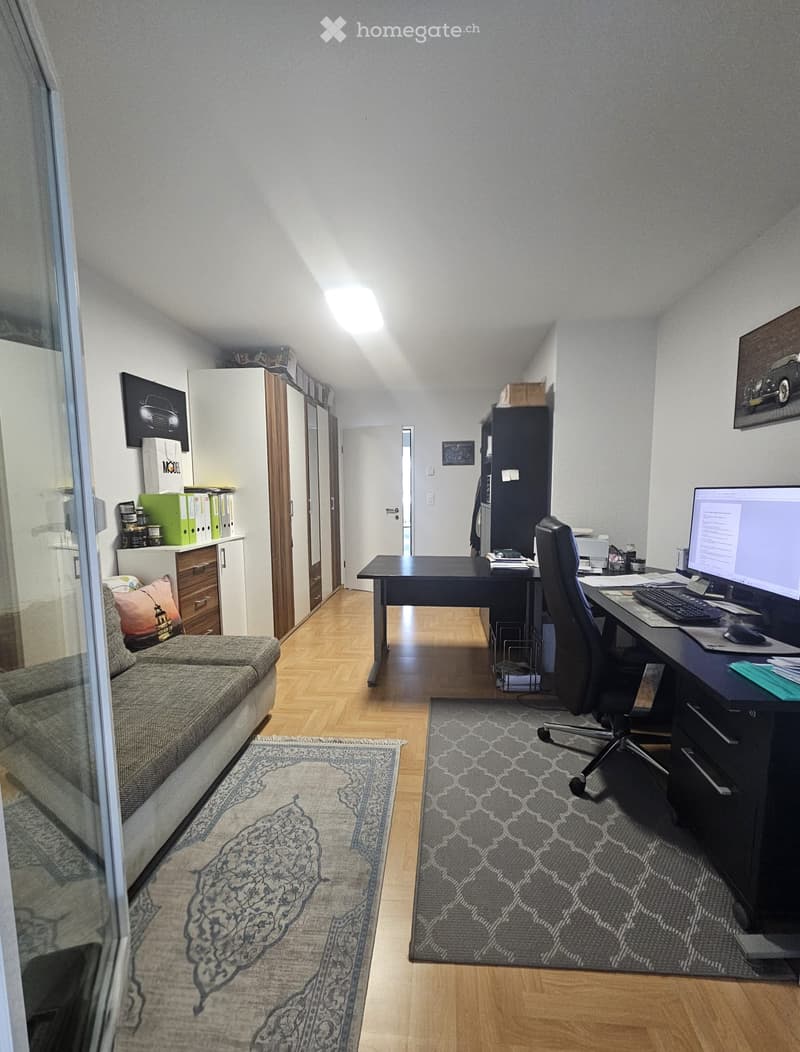 A well-furnished room with a sofa, desk, computer, and various storage units. The room features wooden flooring and multiple rugs.