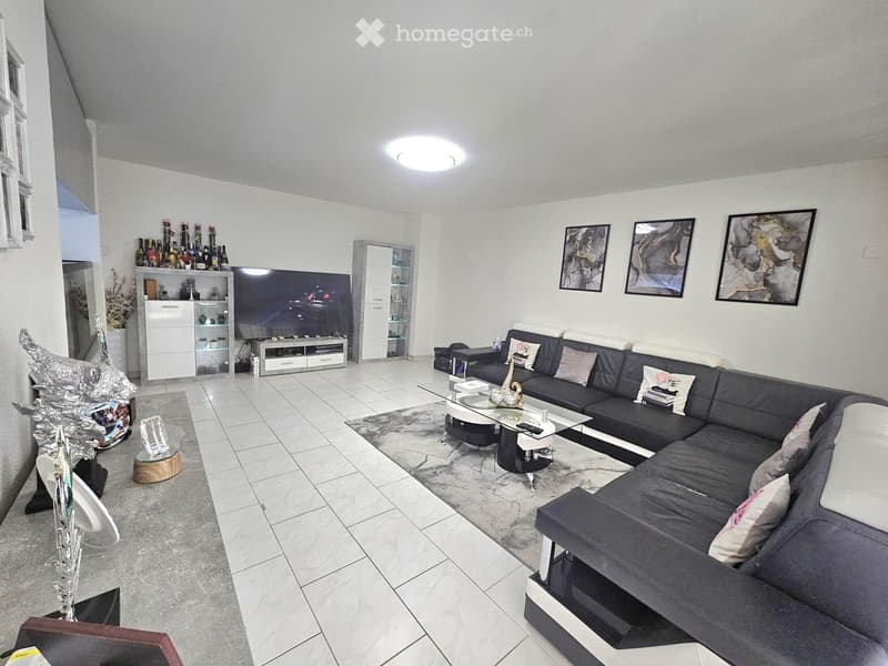 Modern living room with a large sectional sofa, coffee table, TV stand, and decorative shelves. The space features white walls, tiled flooring, and contemporary decor including artwork and a vase.