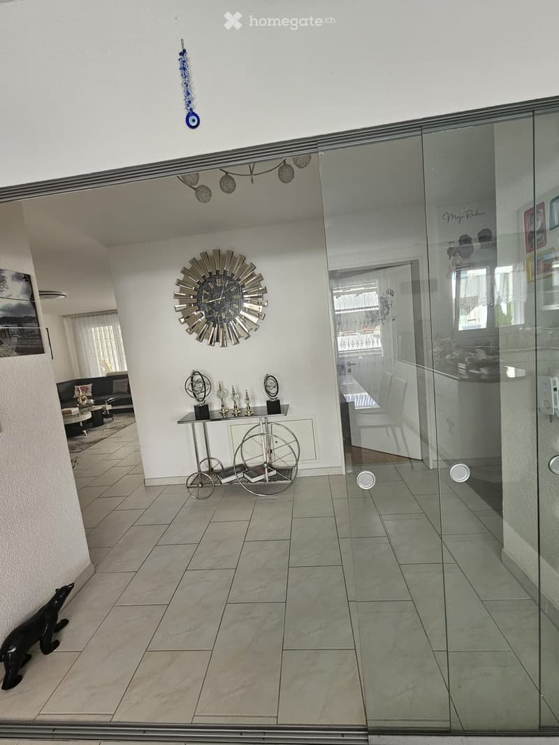Modern hallway with a large glass door leading to another room, featuring a sofa, coffee table, decorative clock, small table with statues, and books on a rolling cart.