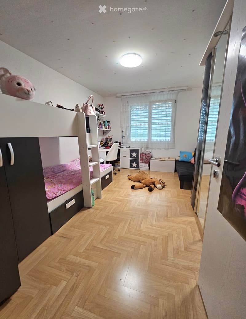 Cozy bedroom with wooden herringbone flooring, bunk bed with pink bedding, desk with chair, shelves, and toys scattered around.