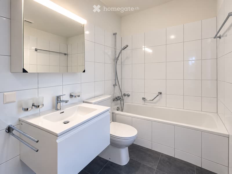 Modern bathroom with white tiles, a sink, toilet, bathtub, and a large mirror above the sink.