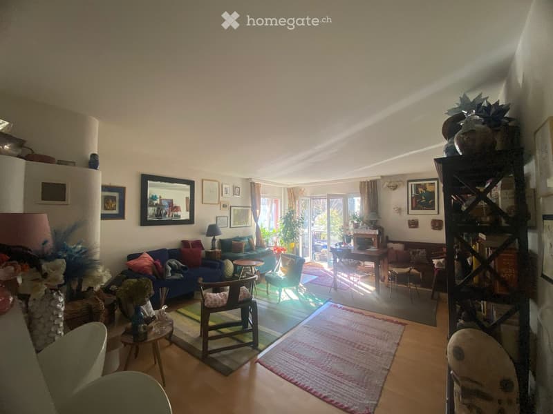 Bright living room with wooden flooring, large windows, and a mix of seating including a blue sofa, armchairs, and a coffee table. Shelves and artwork adorn the walls, and the space is well-lit with natural light.