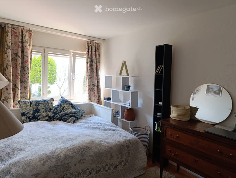 Cozy bedroom with a double bed, pillows, and a blanket. Features a wooden nightstand with a mirror, a shelf with books and decorative items, and large windows with floral curtains.