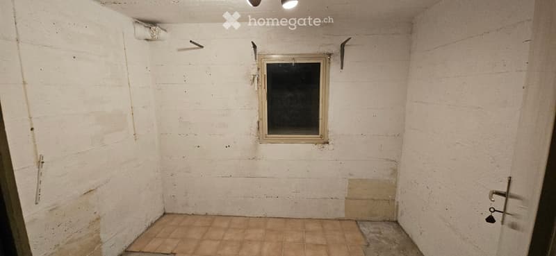 Empty room with bare white walls, tiled floor, small window, and metal hooks on the wall.