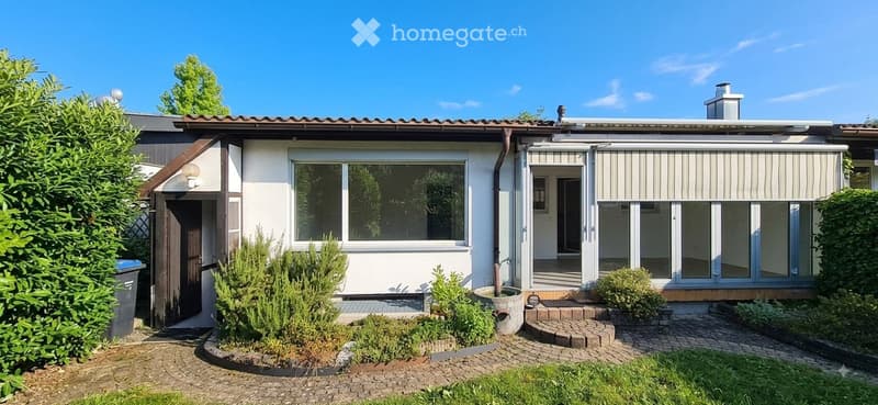 Single-story house with white exterior, large windows, covered entrance, and well-maintained garden.