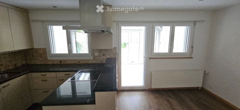 Modern kitchen with light-colored cabinets, dark countertop, range hood, and large windows. Wooden flooring and radiator visible.
