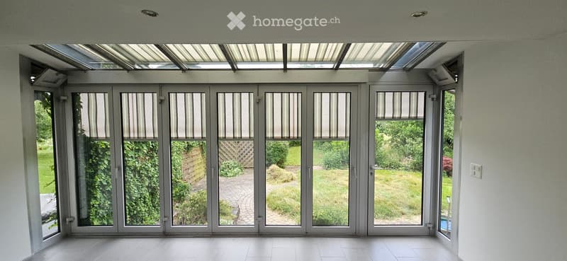 Bright, empty room with large glass doors leading to a garden, white tiled floor, and a modern, minimalist design.