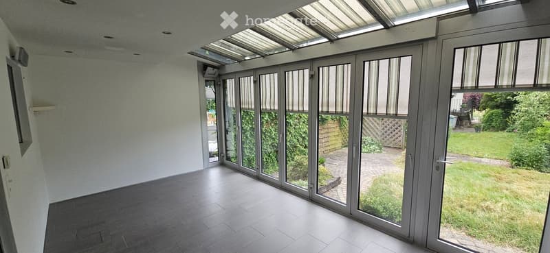 Bright, empty room with large glass doors leading to a garden, tiled floor, and a skylight ceiling.