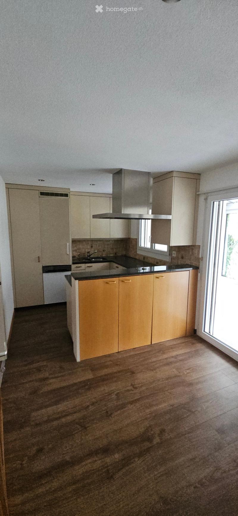 Modern kitchen with light-colored cabinets, a central island, and a black countertop. The floor is made of polished wood, and there is a large window letting in natural light.