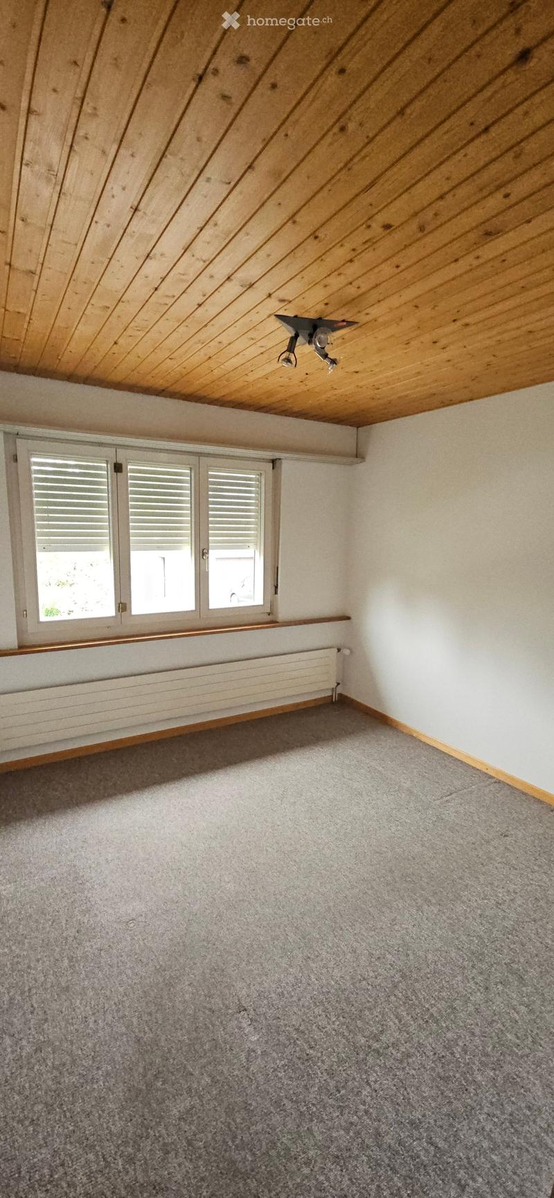 Empty room with grey carpet, white walls, wooden slanted ceiling, and large windows with shutters. No furniture or equipment present.