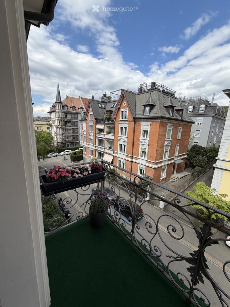 Balcony with green artificial turf, black wrought-iron railing, and potted plants, overlooking a street with historic buildings and parked cars.