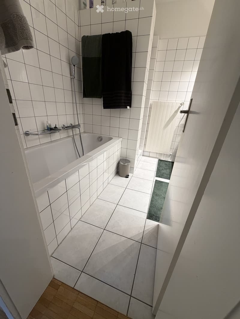 Modern bathroom with white tiled walls and floor, a bathtub, a towel rack with towels, and a small trash can.