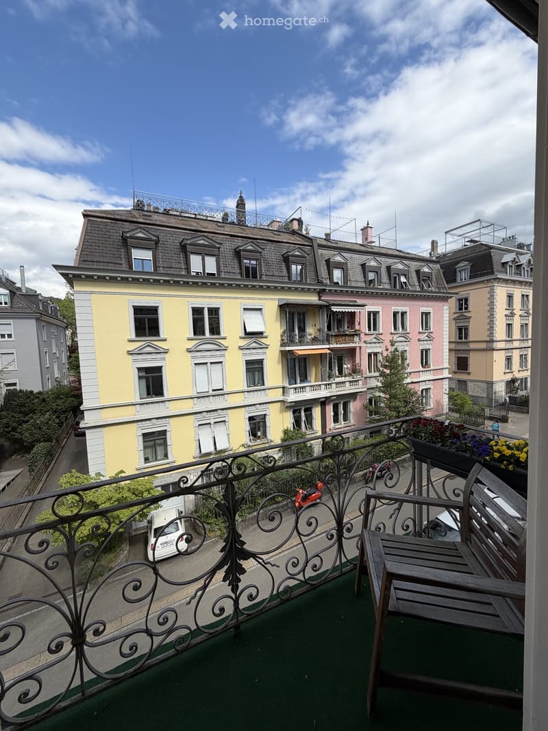 Balcony with green artificial turf, wooden bench, metal railing, and potted flowers overlooking a street with colorful buildings and parked cars.