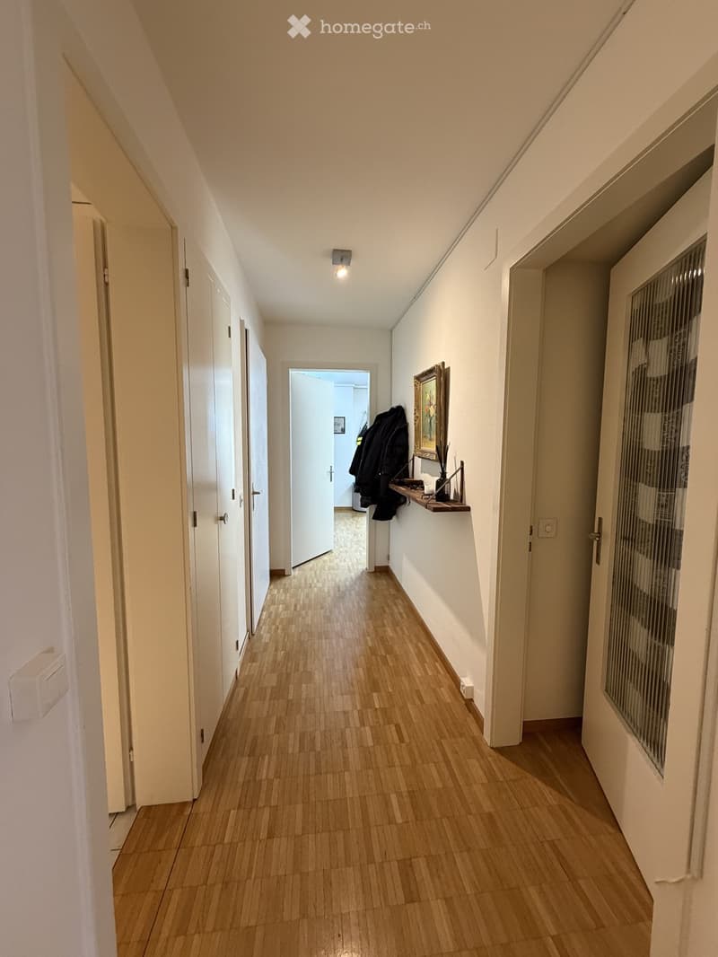 A narrow hallway with wooden flooring, white walls, and several doors. A small shelf with a framed picture and a coat is visible on the right side.