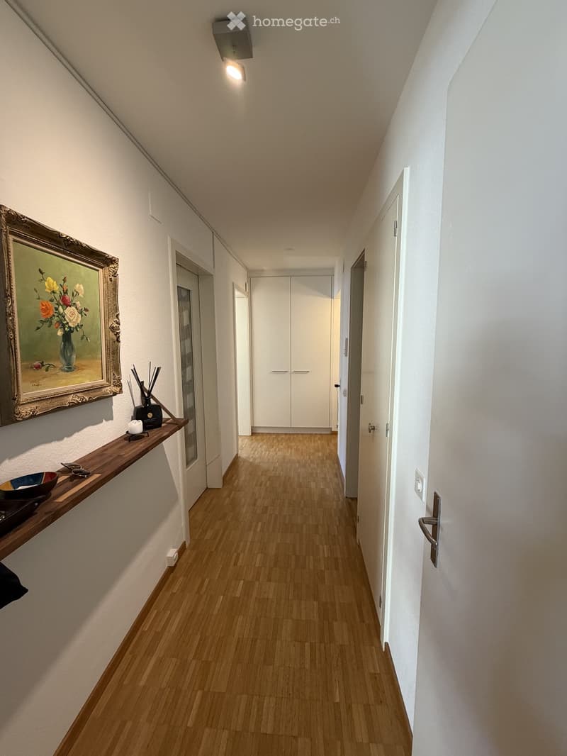 A modern hallway with light wooden flooring, white walls, and built-in white closet units. A small wooden shelf on the left holds decorative items, including a framed painting, candles, and a small bowl.