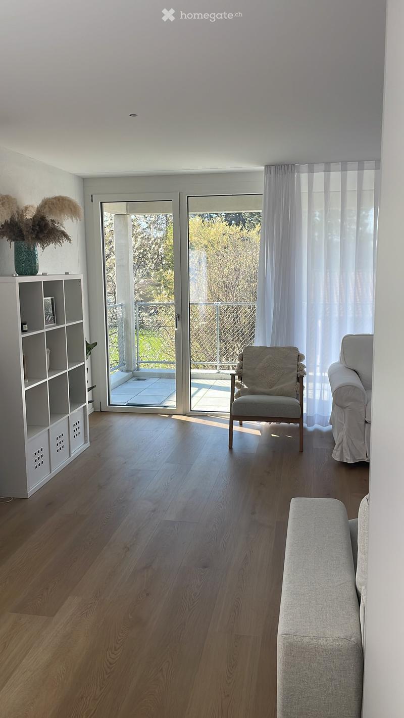 Bright living room with wooden flooring, large sliding glass doors leading to a balcony, a sofa, a chair, and a decorative shelf with a vase.