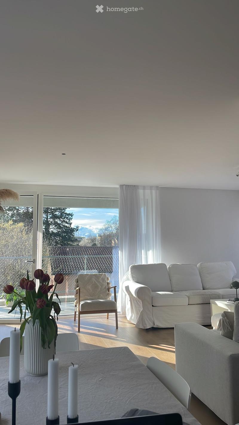 Bright living room with white walls, wooden floor, large windows, sofa, armchair, coffee table, and decorative vase with flowers.