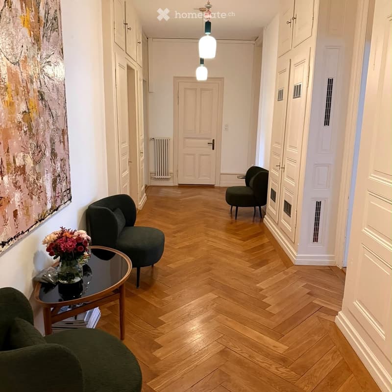 A modern hallway with wooden herringbone flooring, white walls, and several doors leading to other rooms. There are two dark green armchairs, a small round table with a vase of flowers, and a painting on the wall.